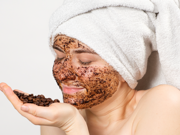 An image of a woman with a coffee grounds face mask and roasted coffee beans in her palm.