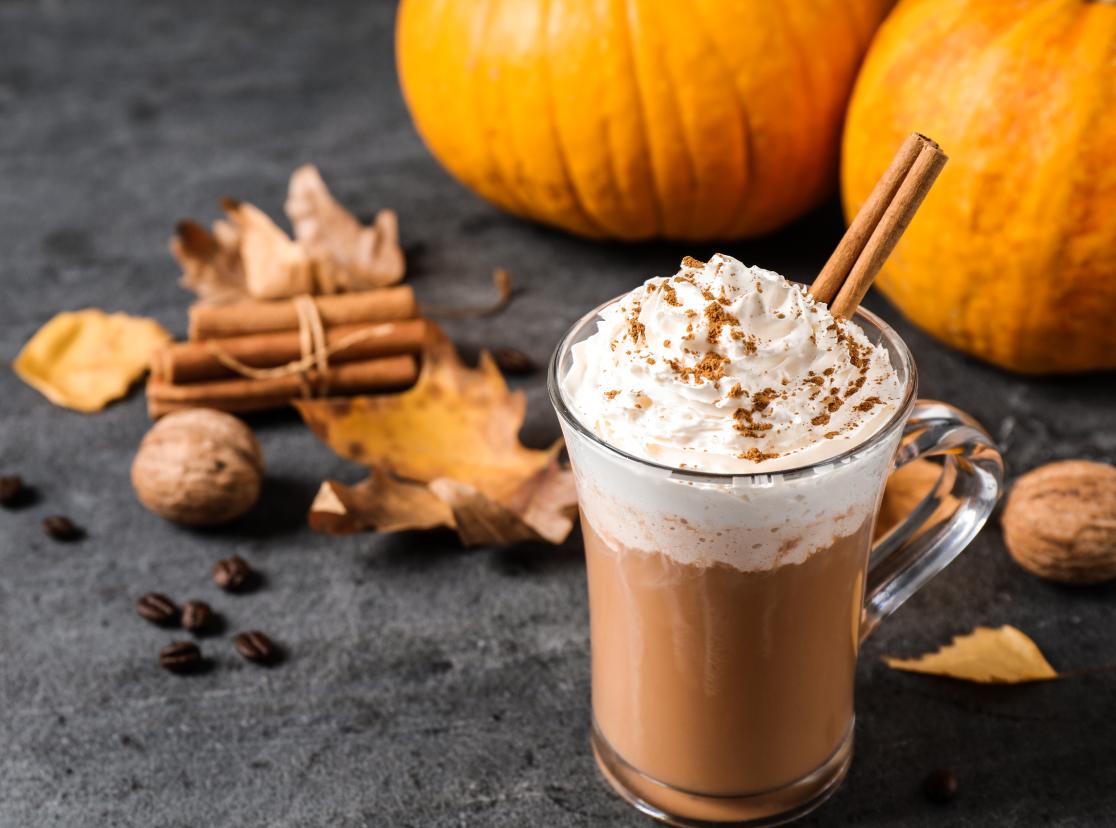 A cup of pumpkin spice latte topped with cream and cinnamon sticks is on a table with A pumpkin sitting behind. Cinnamon, coffee beans, dried leaves, and spices spilled on the table.