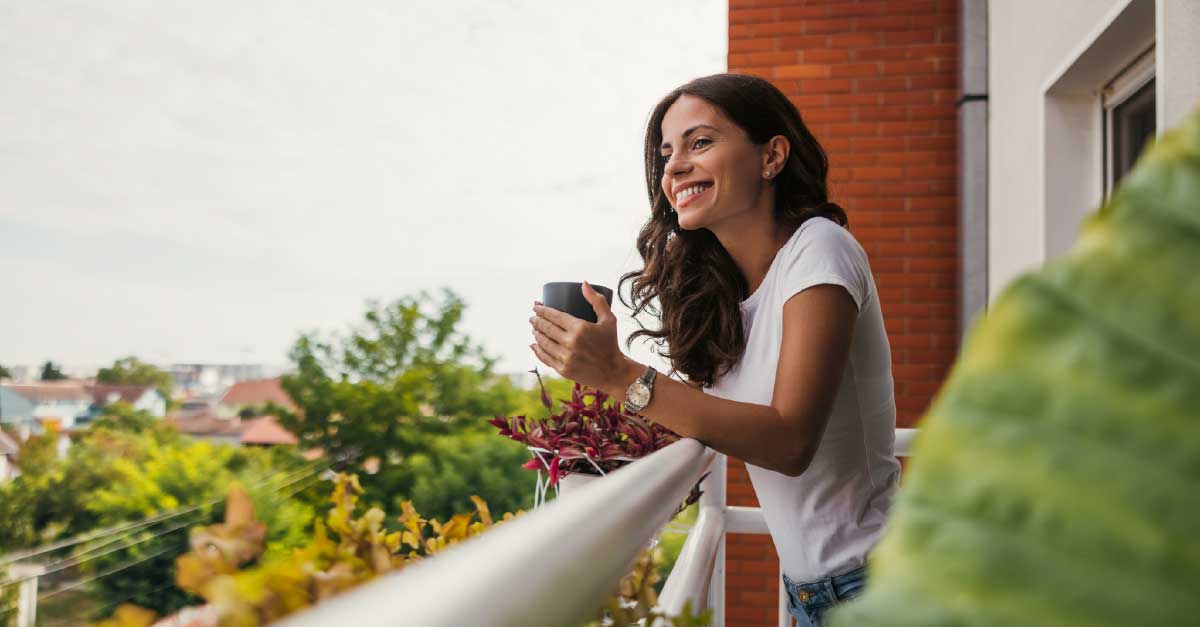 An image of a female drinking mushroom coffee while standing on the balcony of her house.