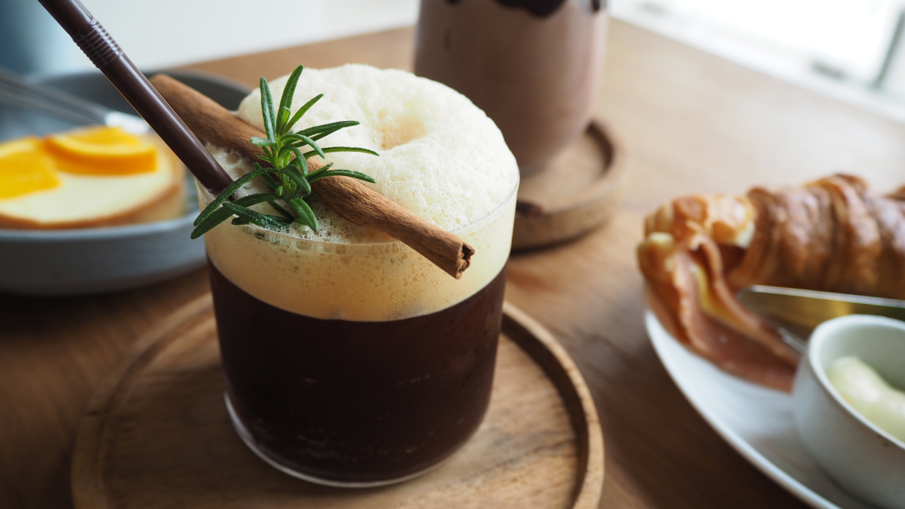 An image of a clear glass filled with Nitro Cold Brew topped with a layer of fizzy foam. Cinnamon sticks and fresh rosemary herbs rest on the foam, adding a touch of aromatic elegance to the beverage.