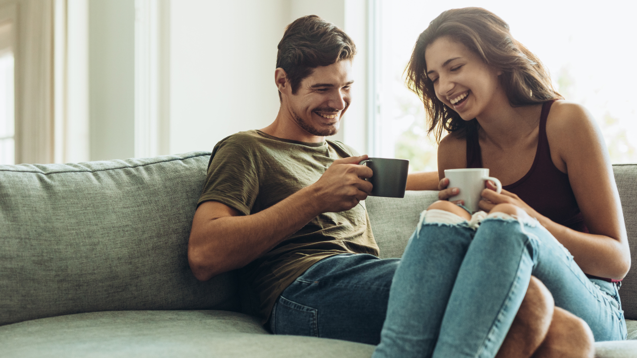 A happy couple sits closely on a cozy sofa, holding specialty coffee cups in their hands, laughing and enjoying a moment of connection. 
