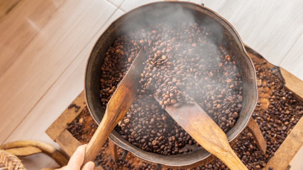 Skillet coffee roasting in action, with coffee beans being stirred in a cast iron skillet over a gas burner, showing the hands-on process of home coffee roasting