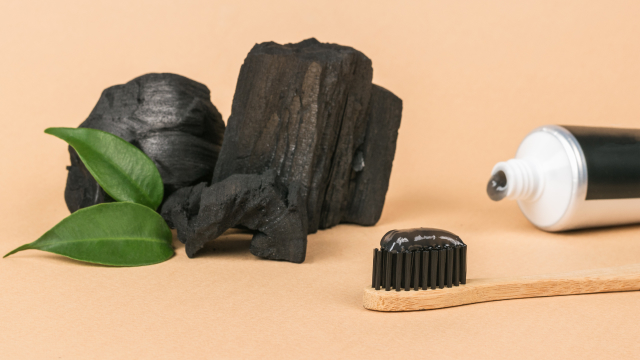 The image features a tube of activated charcoal toothpaste with a dollop on a toothbrush. Against a piece of charcoal, it highlights the product's ability to brighten coffee-stained teeth.