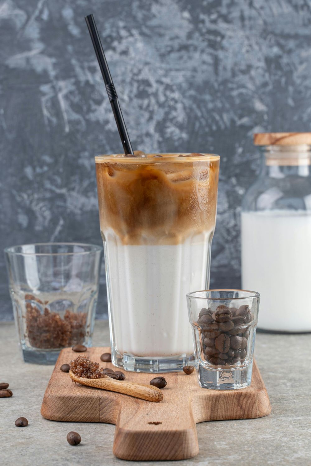 A glass of iced coffee in preparation sits on the table, surrounded by sweetener, roasted coffee beans, and plant-based milk.
