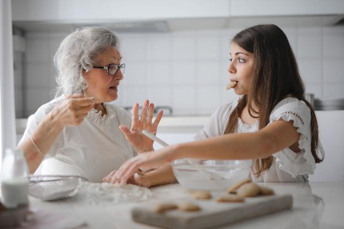 An image captures a heartwarming moment as a smiling grandmother and her young grandchild share a plate of freshly baked cookies, seated at a cozy kitchen table.