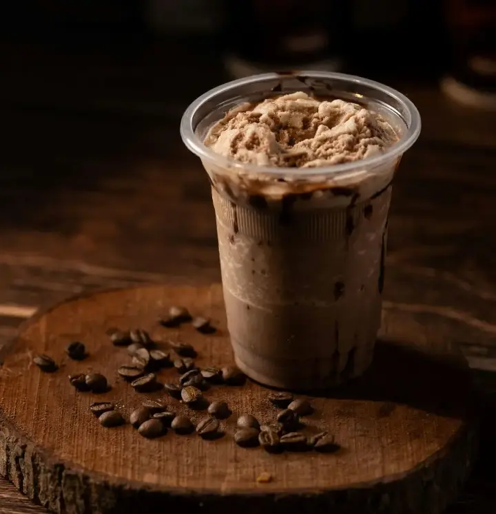 A plastic cup filled with iced coffee mixed with chocolate syrup. Coffee beans are scattered on a wooden coaster, adding a rustic touch to this refreshing iced mocha drink.