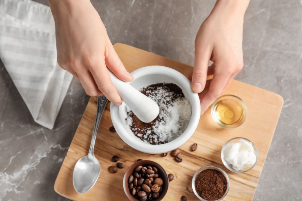 Hands holding a pestle grind coffee grounds and coconut oil in a mortar. Various ingredients, including coffee beans, cinnamon, coconut oil, and sweetener, are ready to create coconut in coffee brew