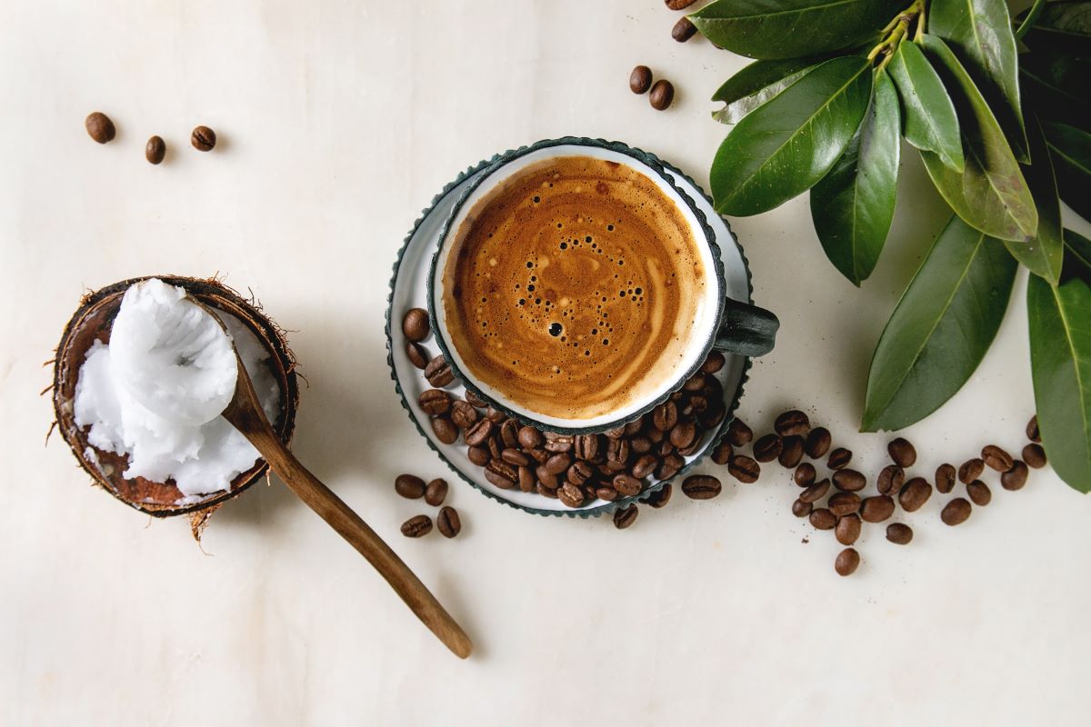 A cup of coffee with a rich crema sits on a saucer surrounded by coffee beans next to a spoonful of coconut oil in a coconut shell.