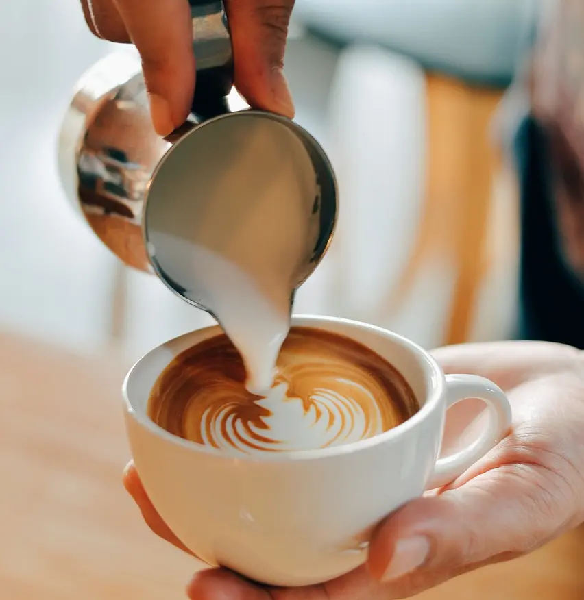 Close-up of a hand pouring steamed milk from a silver pitcher into a cup of espresso, creating intricate cappuccino art on the surface.