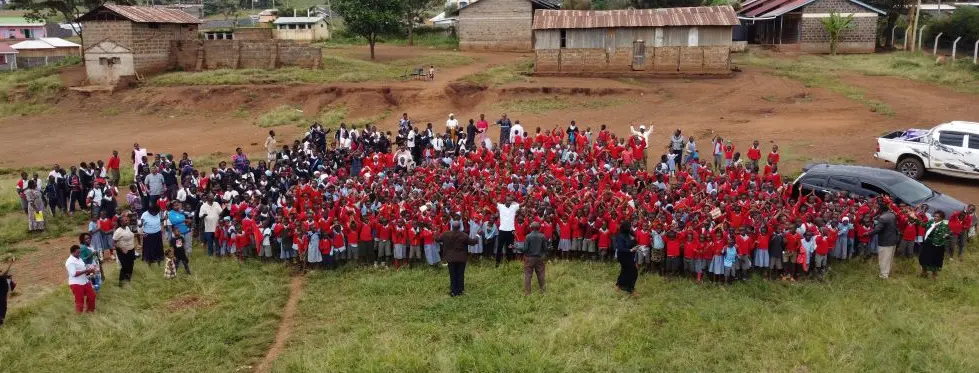 School students and teachers gathered for a photo with the Solai Coffee team. The picture was taken from a drone and shows a wide, open area with a dirt path where floodwaters once swept through. In the background, a single building remains standing, a reminder of the devastating flood that hit the area.