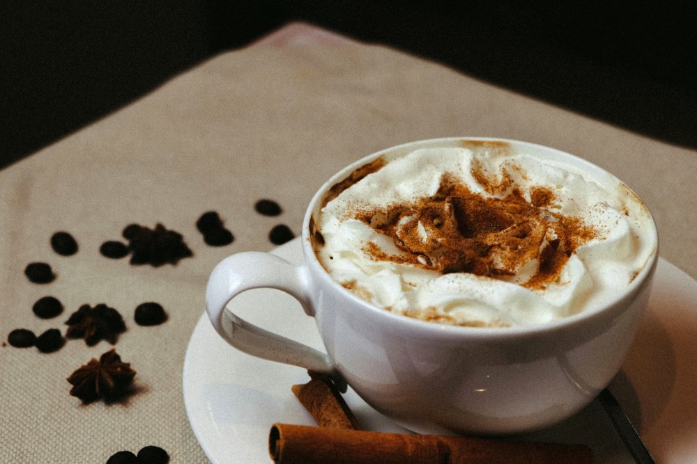 A white ceramic cup of coffee topped with cinnamon-sprinkled milk foam, accompanied by coffee beans and a star anise for a cozy, spiced aesthetic.
