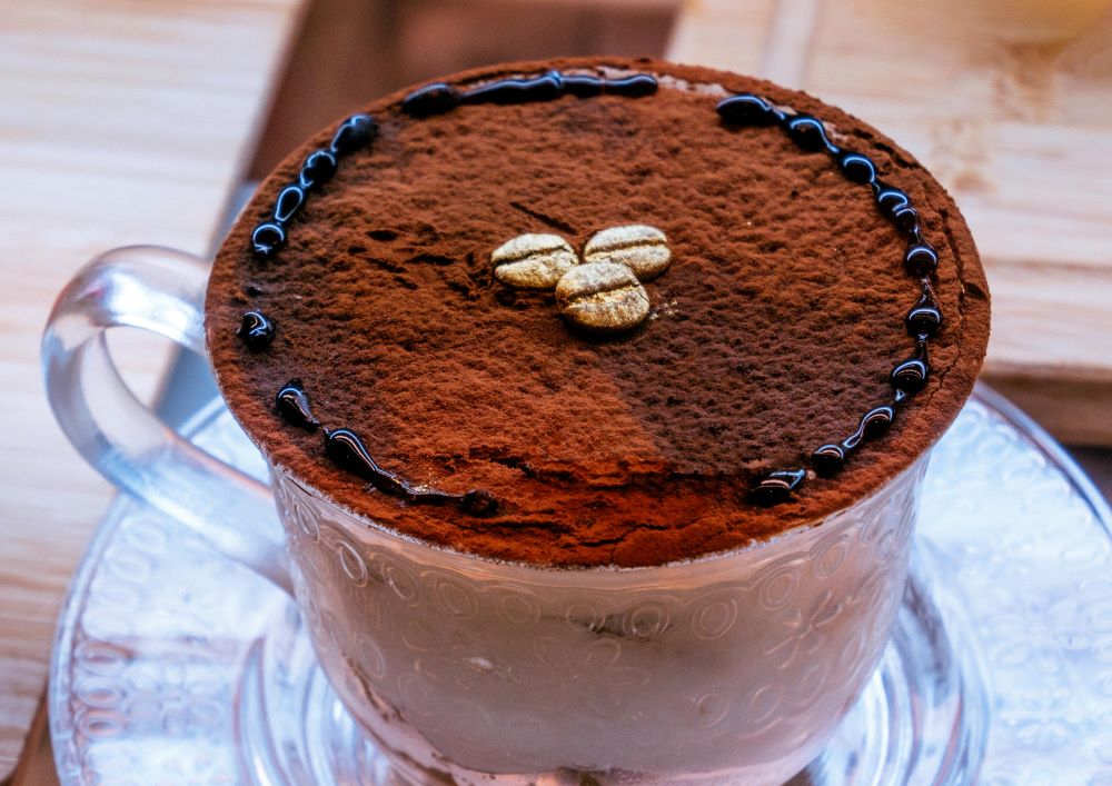 A glass cup filled with a cocoa-infused coffee drink, generously dusted with cocoa powder, surrounded by coffee beans for a rich, indulgent appeal.