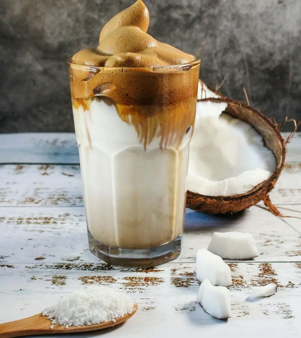 A glass of coffee blended with coconut, combined with coconut flakes and a half coconut shell revealing fresh coconut flesh.