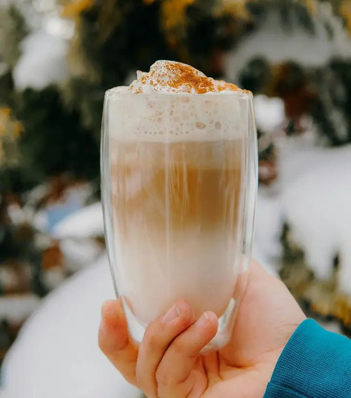 A hand holding a latte in a festive mug, topped with whipped cream and a sprinkle of cinnamon, evoking the warm feeling.