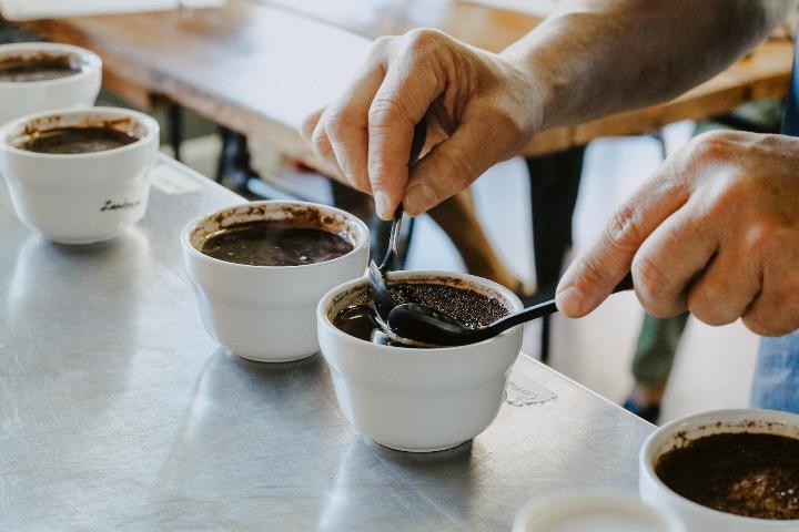 A person performs coffee cupping at home, using two spoons to break the crust on a freshly brewed coffee sample. Several white ceramic cups filled with coffee are on the table.