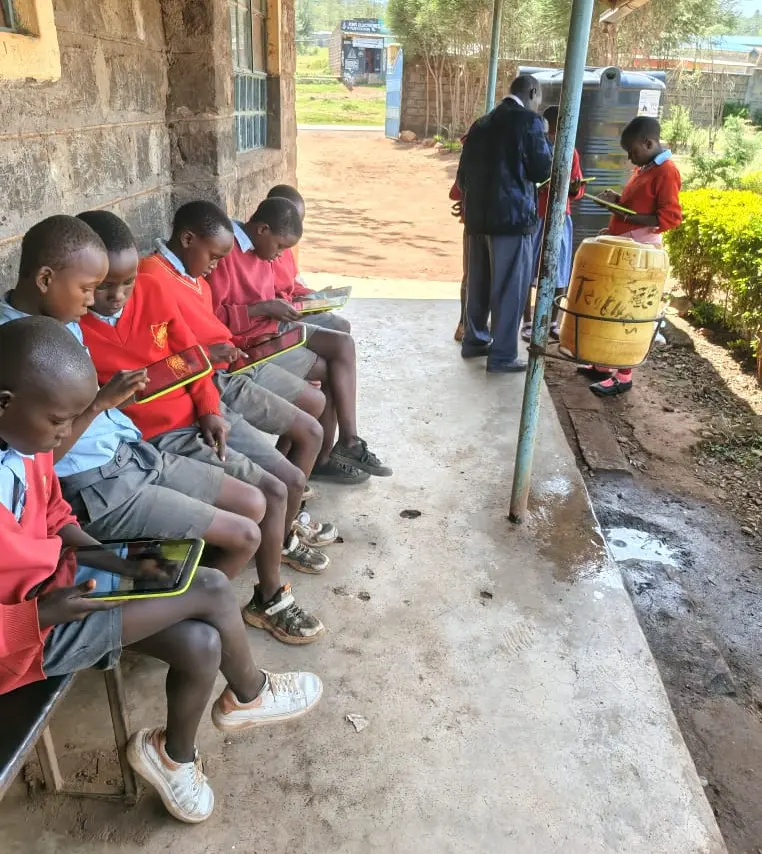 Students at Nyakinyua Primary School enjoy browsing on their tablets outside their classrooms, made possible by the Wi-Fi installation provided by Acts of Wisdom, enhancing digital learning opportunities.
