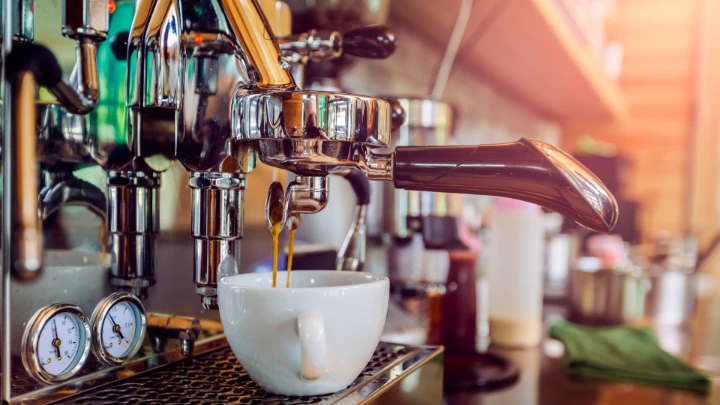Espresso being brewed from a commercial café espresso machine into a white ceramic cup with warm café lighting in the background.