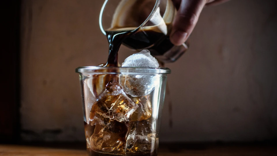 A person pouring cold brew concentrate into a glass filled with ice before adding cold water and milk, with the concentrate bottle visible in the background.