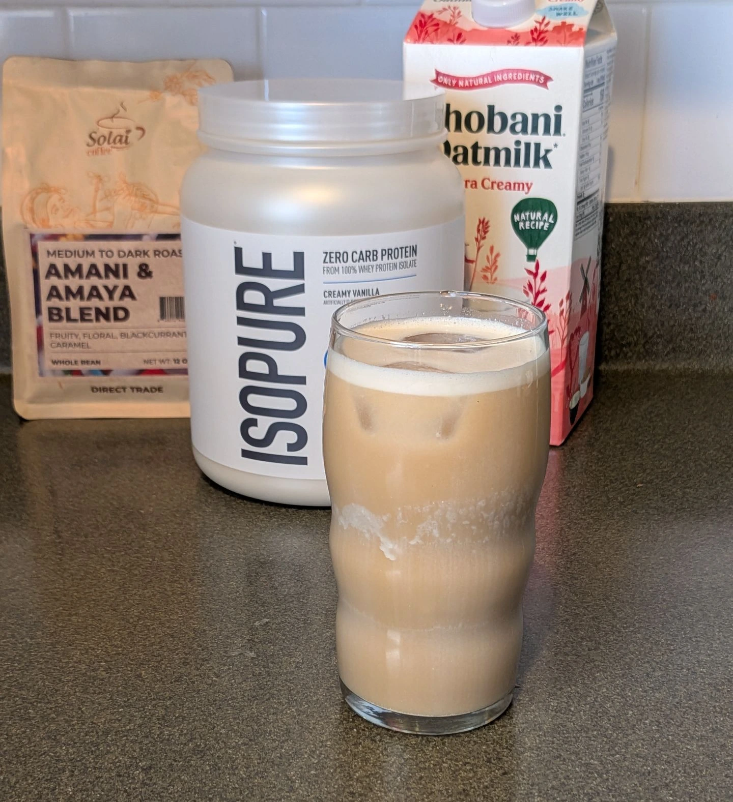 Glass of iced protein coffee made with Solai cold brew, vanilla Isopure protein powder, and Chobani oat milk alongside the ingredients on a kitchen countertop.