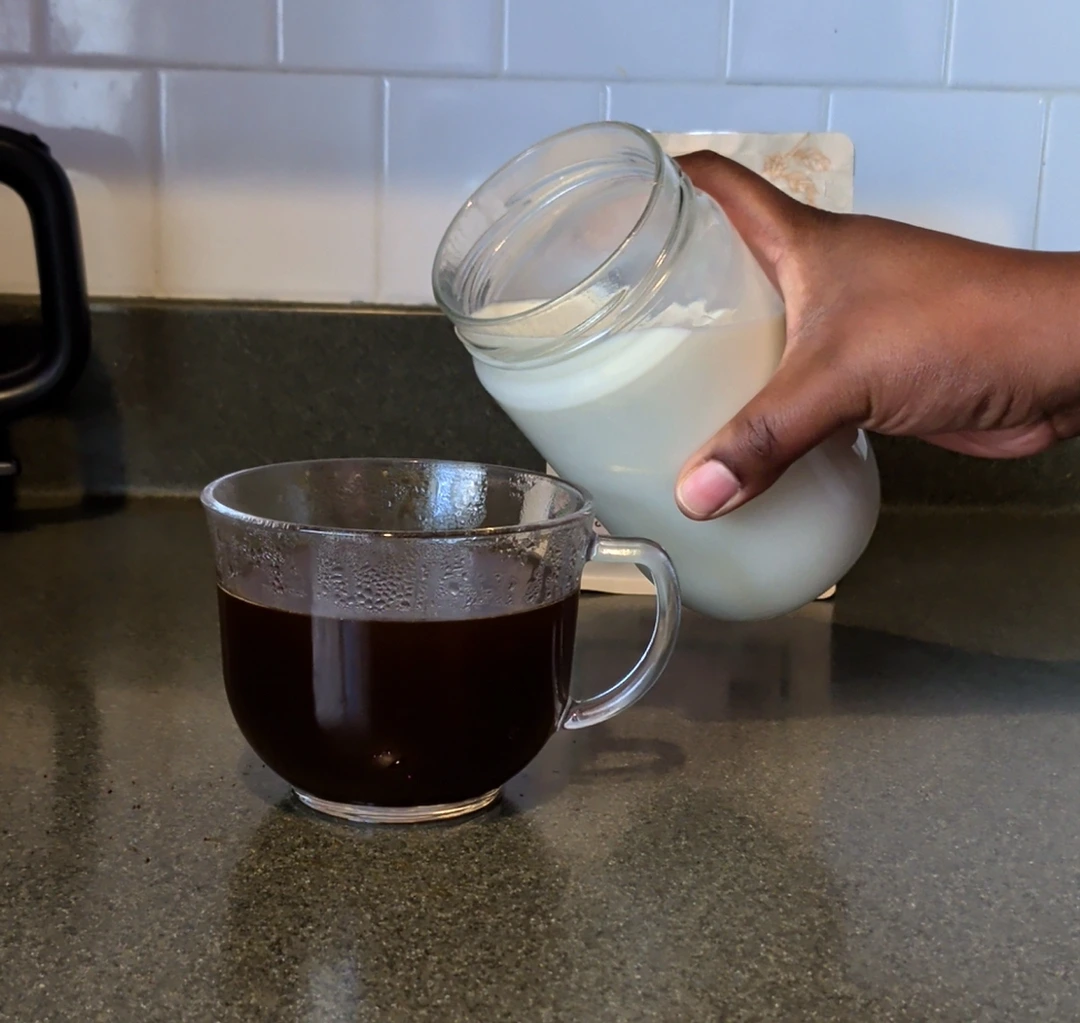 Hand pouring a vanilla protein and oat milk mixture into a cup of freshly brewed Solai coffee before stirring to make homemade protein coffee.